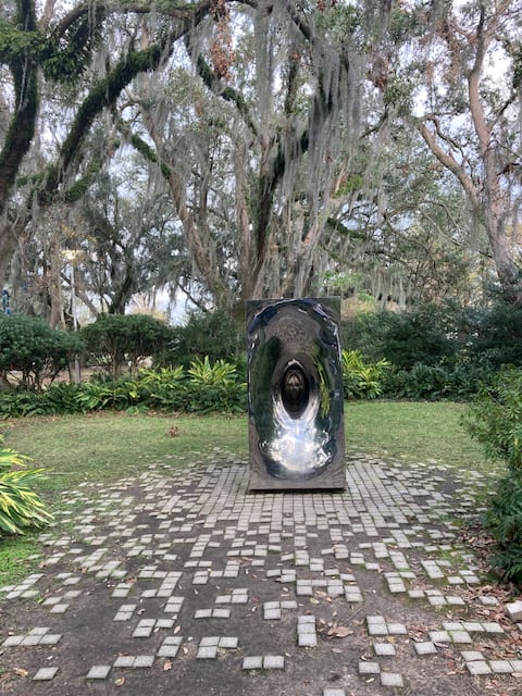 A concave shiny steel sculpture in front of moss-draped live oaks