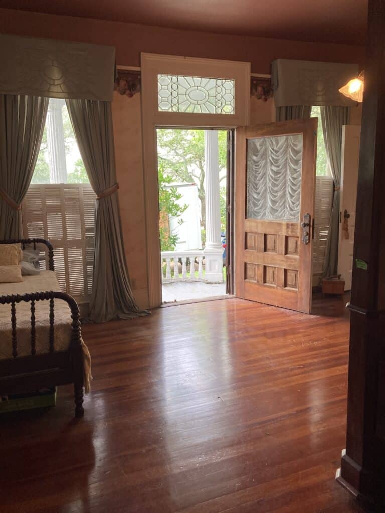 An old-fashioned bedroom with the door to a side porch open. Elaborate transom window, hardwood floors, and puddling blue silk drapes. No watermelon present. 