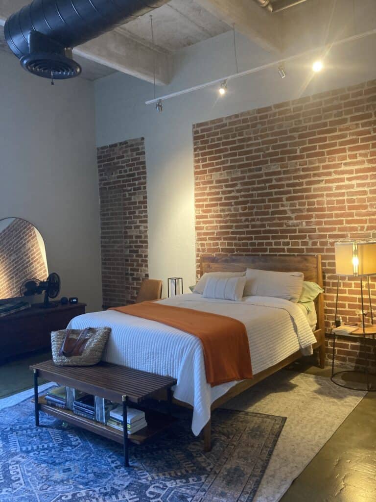 A bedroom that generates a feeling of contentment with exposed brick, a white bedspread, blue overlaid rugs, and a slat bench at the foot of the bed.