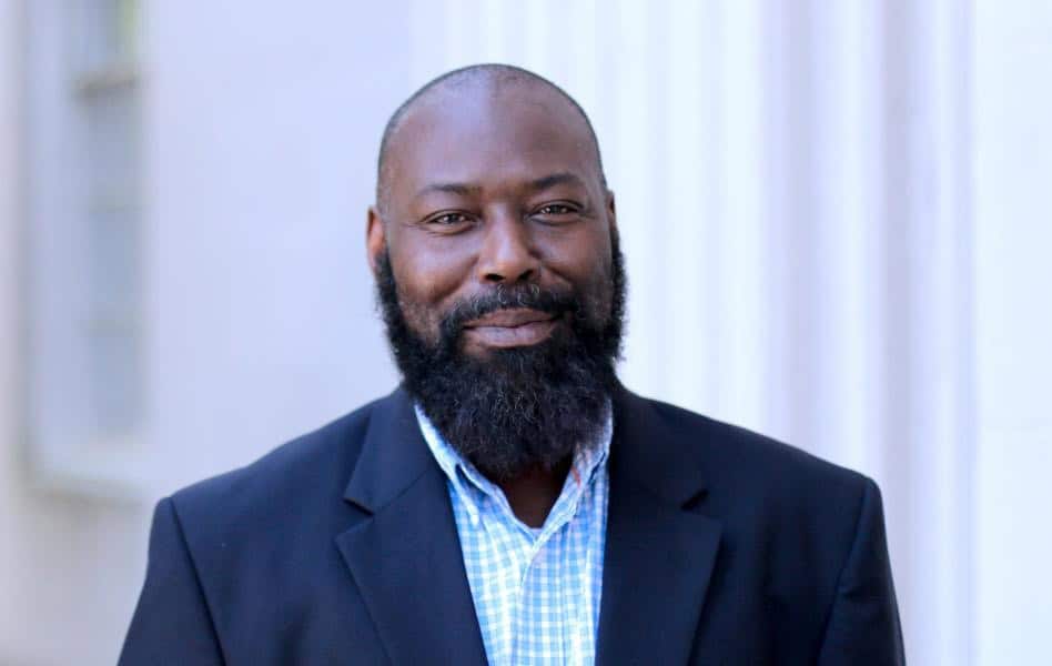 Head shot of Black man Randall Kenan with a vee shaped beard, smiling, in a navy jacket and light blue check shirt. 