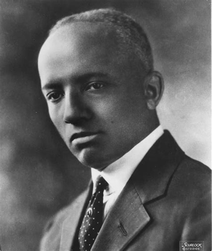 Portrait of Carter Woodson, who created the origin of Black History Month. He appears middle aged, with a high forehead and wearing a dark suit, white shirt, and dotted tie with what appears to be a fraternity tie clasp. 