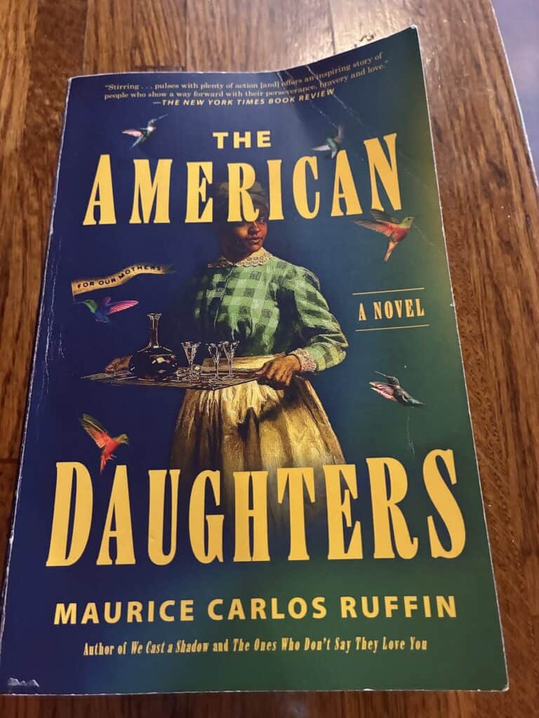 Photo of the cover of MarcusCarlos Ruffin's novel The American Daughters with a navy background and yellow font on the title and a photo of a Black woman in a lace collar, gingham shirt, and white pleated apron, carrying a tray.