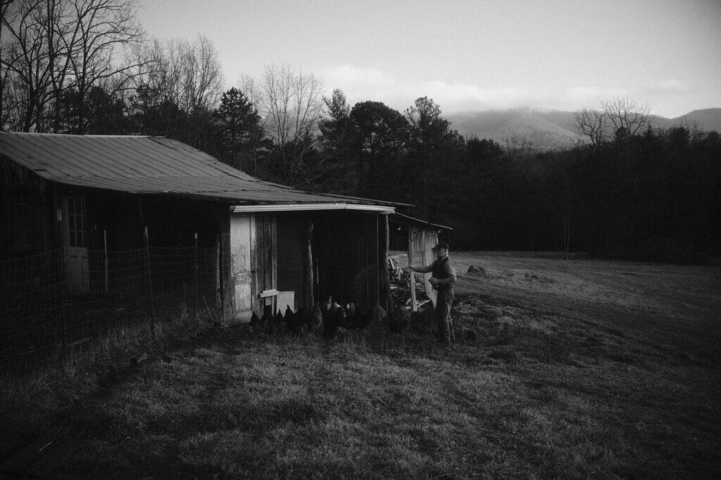 A black and white photo of a man opening the porch door of a tin-roofed cabin with mountains in the distance