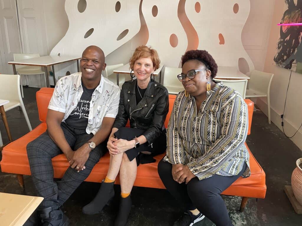 Maurice Ruffin, me, and Desiree Evans on an orange vinyl couch with a modern wavy sculpture on the wall behind us, enjoying the magic of the book launch.